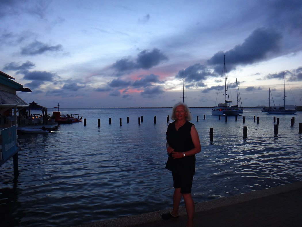 Person standing on waterfront promenade at dusk with sailboats and wooden posts in calm water under a cloudy sky with hints of sunset colors
