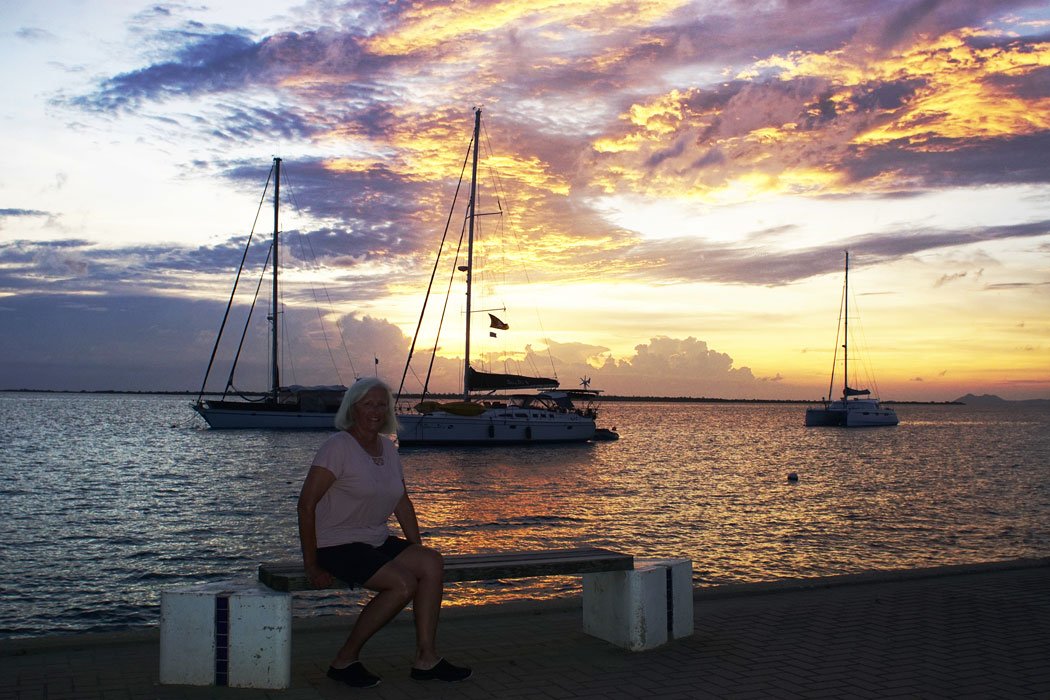 Person sitting on a bench near calm water with sailboats anchored in the background under a colorful sunset sky
