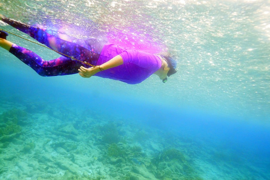 Person snorkeling underwater wearing a purple top and patterned leggings, holding a rope attached to an inflated buoy on the surface