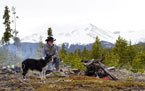 Person sitting on a rock in a mountainous forest clearing with a dog, snow-capped peaks in the background