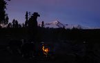Campfire glowing softly under a dark sky with silhouetted trees and distant snow-covered mountain