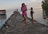 Two people walking on a stone pier extending into calm water with a soft, pastel-colored sky at sunset or sunrise