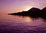 Calm sea at sunset with purple and pink hues reflecting on the water and a dark silhouette of a mountainous coastline