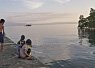 Two people sitting and standing on a rocky shore, looking out at a calm sea with a small island and cloudy sky in the background at sunset