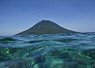 Calm ocean water with gentle waves in the foreground and a conical volcanic island rising in the background under a clear sky