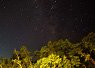 Starry night sky above a tree-covered landscape with hills silhouetted against the horizon