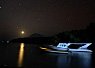 Illuminated boats floating on calm water under a dark night sky with a bright moon and distant hills