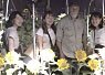 A group of people dressed in light-colored clothing standing closely together, illuminated by surrounding flowers and soft lighting, creating a serene and intimate atmosphere