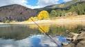 Fishing rod angled over calm river water with autumn trees and forested mountains in the background under a partly cloudy sky