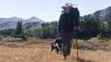 Man wearing a wide-brimmed hat and carrying a large backpack walking with a black and white dog through a dry grassy field with mountains and trees in the background