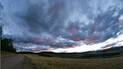 Wide open landscape with a dirt road curving left, grassy fields, forested hills, and a dramatic cloudy sky illuminated by the setting sun