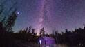 Star-filled night sky with the Milky Way visible above a small cabin surrounded by trees