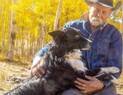 Man wearing a cowboy hat and denim jacket sitting outdoors among yellow autumn aspens, holding and petting a black and white dog on his lap