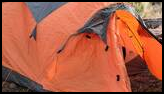 Orange and gray tent pitched on a forest floor with pine needles and pinecones, surrounded by trees and overlooking distant mountains
