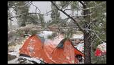 Orange tent partly covered with snow set up among pine trees and rocks in a snowy outdoor setting