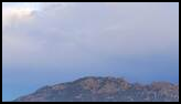 Foggy mountain ridge seen through a frame of tall pine trees under a cloudy sky with patches of blue