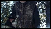 Man wearing a dark cowboy hat and camouflage vest gently petting a black and white dog dressed in a green plaid coat near a small campfire in a pine forest