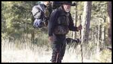 Man wearing a black cowboy hat and carrying a large backpack walking through a snowy forest clearing with two dogs and using a wooden walking stick