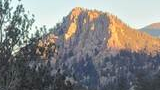 Sunlit rocky mountain peak with surrounding forested hills and trees in the foreground under a clear sky