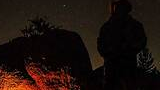 Silhouette of a person wearing a hat standing near glowing campfire rocks under a starry night sky