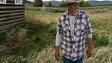 a man in a cowboy hat and plaid shirt walking through tall grass beside a rustic log cabin in a wide open field with mountains in the distance