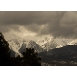Snow-covered mountain peaks beneath a dramatic cloudy sky with dark trees in the foreground