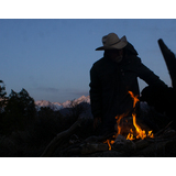 Person in a hat tending a small campfire at dusk with snow-capped mountains and trees in the background