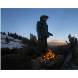 Man wearing a cowboy hat and jacket standing near a small campfire surrounded by rocks with snow-covered mountains and trees in the background at dusk