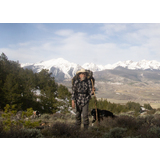 Person wearing a cowboy hat and backpack hiking with two dogs in a mountainous landscape with snow-covered peaks in the background
