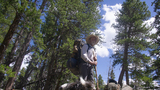 A man wearing hiking gear and a wide-brimmed hat stands on rocky ground surrounded by tall pine trees with a cloudy blue sky above, while a black and white dog is in the foreground