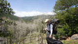 Older person with large backpack and hiking stick petting a black dog on rocky terrain with mountains and leafless trees in the background