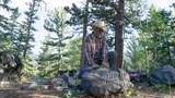 Man wearing a cowboy hat and plaid shirt sitting on forest ground surrounded by camping gear and three black and white dogs resting nearby