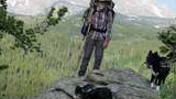 Hiker with backpack and walking stick standing on a rocky outcropping with two black and white dogs, surrounded by pine trees and mountains in the background