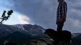 Man wearing a wide-brimmed hat and plaid shirt standing on a rock outcropping with two black and white dogs, overlooking a mountainous landscape under a cloudy sky at dusk