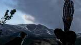 Man in a plaid shirt and cowboy hat standing on a rocky ledge with two black and white dogs, under a cloudy evening sky with snow patches on distant mountains