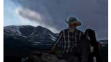 Man in cowboy hat and plaid shirt sitting on rocks with two black and white dogs, snowy mountain and cloudy sky in the background at dusk
