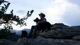 man wearing a cowboy hat sitting on a large rock outcropping with two black and white dogs, with a backdrop of pines and snow-capped mountains under a cloudy sky at dusk