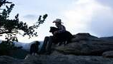 Person wearing a wide-brimmed hat sitting on rocks with two black and white dogs, overlooking a mountainous landscape during early evening