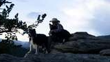 Person wearing a wide-brimmed hat sitting on rocky outcropping hugging one of two black and white dogs, with pine trees and snow-capped mountains in the background at dusk