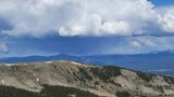 Expansive mountain landscape with a ridge in the foreground, patches of snow, dense evergreen forest below, and distant mountain ranges under a sky filled with large, dramatic clouds