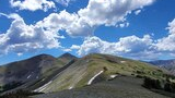 Mountain ridge under a blue sky filled with scattered white clouds, with patches of snow on the slopes and sparse vegetation