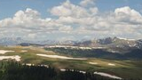 Expansive alpine landscape with patches of snow on rolling green hills, dense spruce fir forest below, and distant snow-capped mountain peaks under a blue sky with scattered clouds