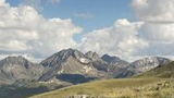Rocky mountain peaks with patches of snow under a blue sky filled with large, fluffy white clouds, overlooking a grassy alpine meadow with scattered rocks and wildflowers