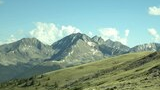 Mountain range with rocky peaks and patches of snow under a blue sky with scattered clouds, viewed from a grassy hillside with scattered rocks and a few small trees