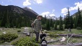 Man in a plaid shirt and hat standing in a forested mountain clearing with two black and white dogs, surrounded by evergreen trees and rocky peaks under a partly cloudy sky