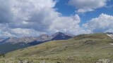 Expansive alpine meadow with wildflowers in the foreground and rugged mountain peaks under a sky filled with fluffy clouds