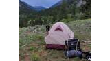 Small tent pitched on a grassy mountain valley with two border collie dogs nearby and forested mountains in the background under a cloudy sky