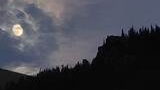Bright full moon partially obscured by thin clouds over the silhouette of a forested mountain ridge at dusk