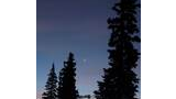 Crescent moon and a bright planet visible in the early evening sky above dark silhouettes of tall pine trees with a mountain ridge in the background