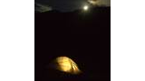 Illuminated tent glowing warmly in the dark outdoors with a bright moon rising over a forested mountain ridge